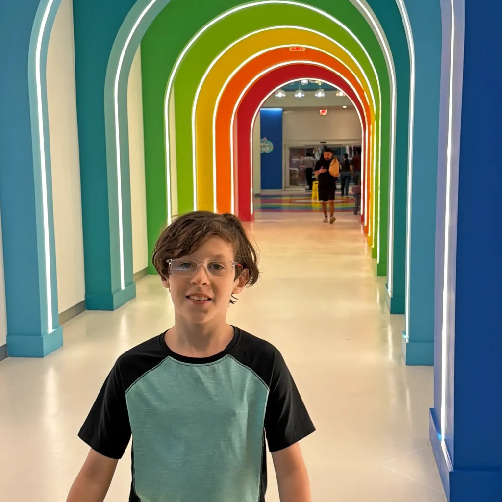a Boy with Glasses Stands Smiling in Front of a Bright Hallway with Rainbow-colored Illuminated Arches. Two People Are Walking in the Background Through the Colorful Passage at Universal Kids Resort in Frisco, Texas. - Favorite Grampy Travels - Favorite Grampy Travels 2026