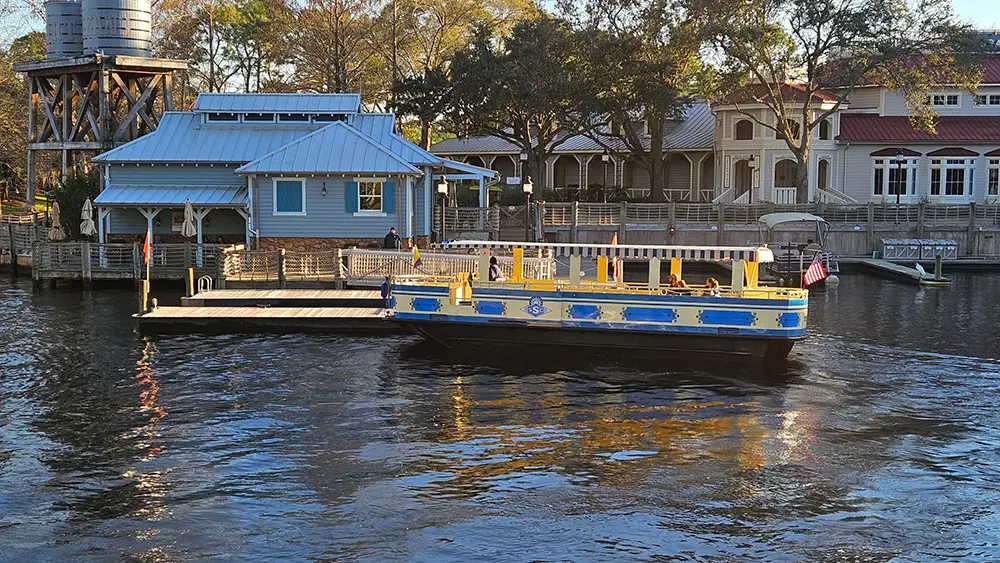 Wdw-port-orleans-riverside-boat-ferry-transportation - Favorite Grampy Travels a Blue and Yellow Ferry Boat is Docked by a Wooden Pier Near a Light Blue Building with a Metal Roof, Reminiscent of the Charming Port Orleans Riverside Resort at Disney. Trees and Other Buildings Are Visible in the Background on a Sunny Day. - Favorite Grampy Travels - Favorite Grampy Travels 2026