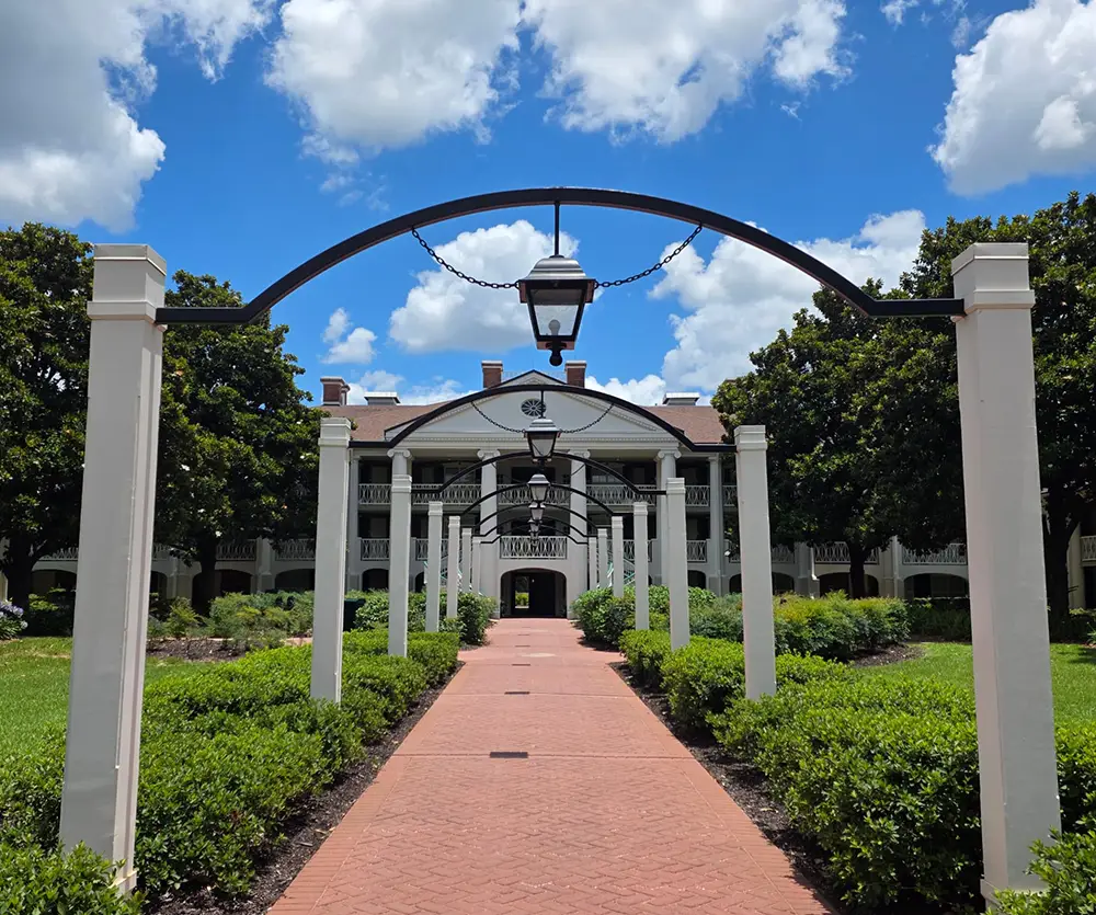 Wdw-port-orleans-riverside-magnolia-bend-pathway - Favorite Grampy Travels a Brick Pathway Leads Through Arches and Greenery to a Large Building with White Columns, Black Trim, and a Central Lantern Under a Blue Sky with White Clouds, Evoking the Elegant Charm of Disney’s Port Orleans Riverside Resort. - Favorite Grampy Travels - Favorite Grampy Travels 2026