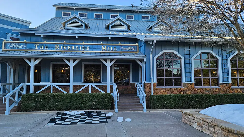 Wdw-port-orleans-riverside-the-riverside-mall - Favorite Grampy Travels Blue Building with a Sign Reading the Riverside Mill, White Railing, Large Windows, and a Giant Outdoor Chessboard Set Up on the Pavement in Front—this Charming Spot is Part of Disney’s Port Orleans Riverside Resort. - Favorite Grampy Travels - Favorite Grampy Travels 2026