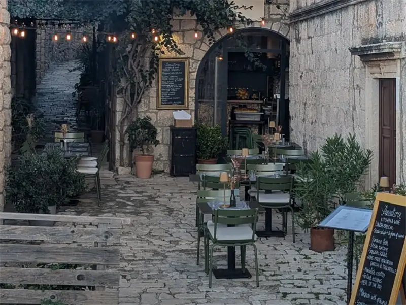 Outdoor Café with Green Chairs and Tables on a Cobblestone Street, Surrounded by Stone Buildings, Potted Plants, String Lights, and Menu Boards. the Setting Appears Cozy and Inviting. - Favorite Grampy Travels - Favorite Grampy Travels 2026 - Favorite Grampy Travels 2026