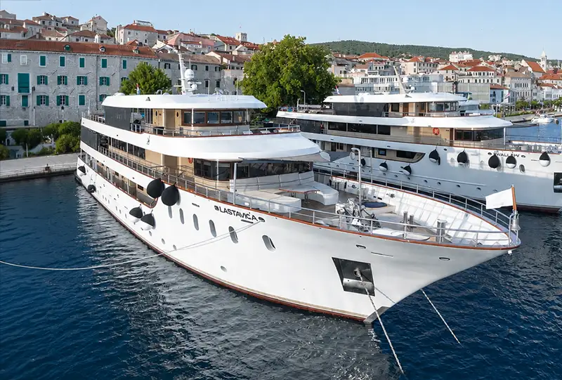 a Large White Yacht Named Lastavica is Docked in a Harbor in Croatia, with Her Sister Ship Nearby. Behind Them Are Waterfront Buildings with Red Roofs and a Hillside Covered in Greenery. the Sea is Calm Under a Clear Sky. - Favorite Grampy Travels - Lastavica Cruising - Favorite Grampy Travels 2026 - Favorite Grampy Travels 2026