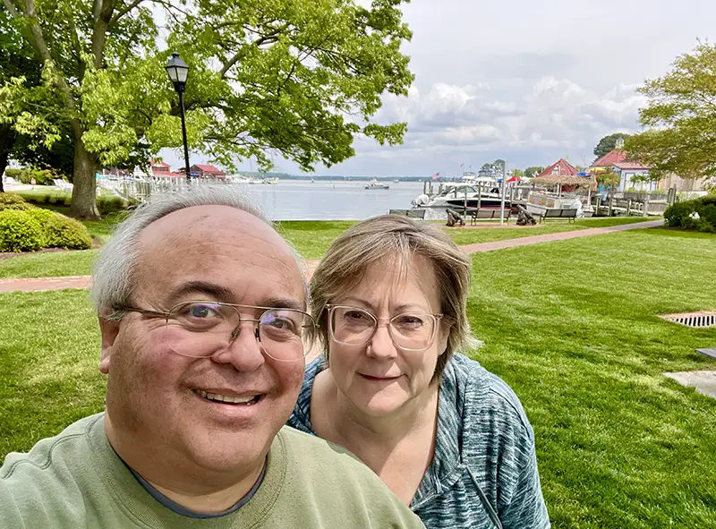 a Man and Woman Smile for a Selfie in a Park with Green Grass, Trees, and Boats Docked by a Lake in the Background on a Cloudy Day. - Favorite Grampy Travels - Favorite Grampy Travels 2026 - Favorite Grampy Travels 2026