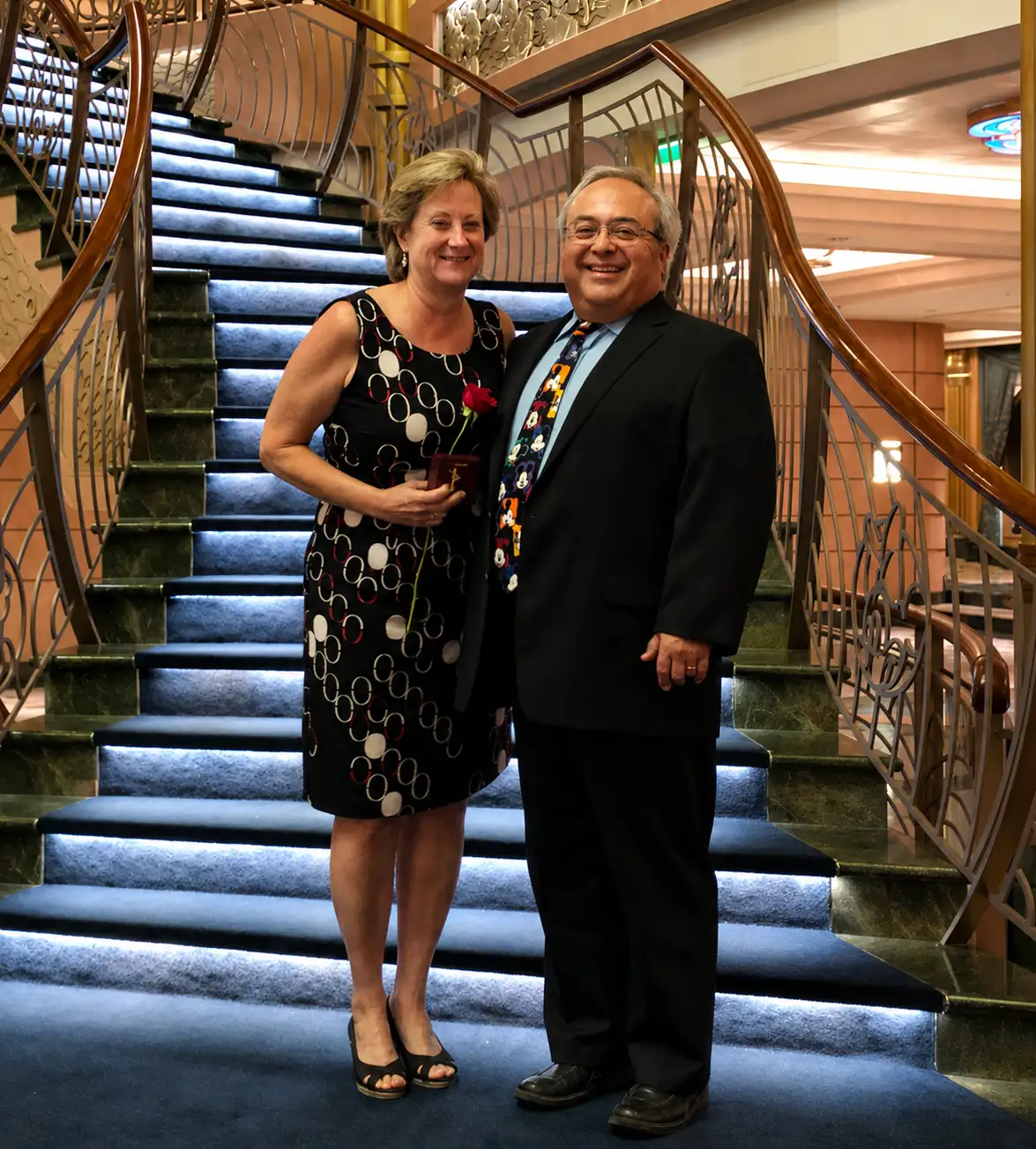Grammy Holding a Red Rose and Favorite Grampy in a Suit Stand Smiling on a Staircase with Blue-lit Steps and Ornate Railings in an Elegant Indoor Setting. - Favorite Grampy Travels - Favorite Grampy Travels 2026 - Favorite Grampy Travels 2026