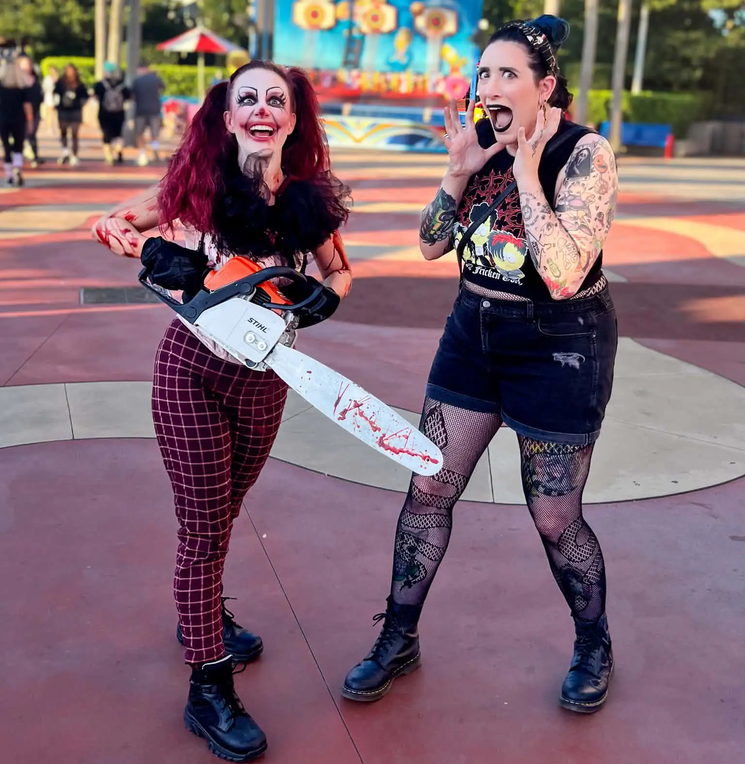 Two women pose outdoors; one wears clown makeup and holds a bloody chainsaw, smiling. The other, covered in tattoos and gothic attire, looks shocked with hands near her face. It appears to be a playful, staged scene. - Favorite Grampy Travels