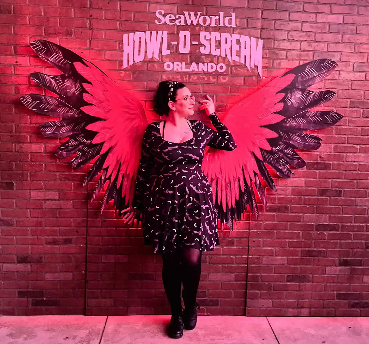 A person in a black dress poses in front of a brick wall with red and black wings and a SeaWorld Howl-O-Scream Orlando sign, creating the illusion they have wings. - Favorite Grampy Travels