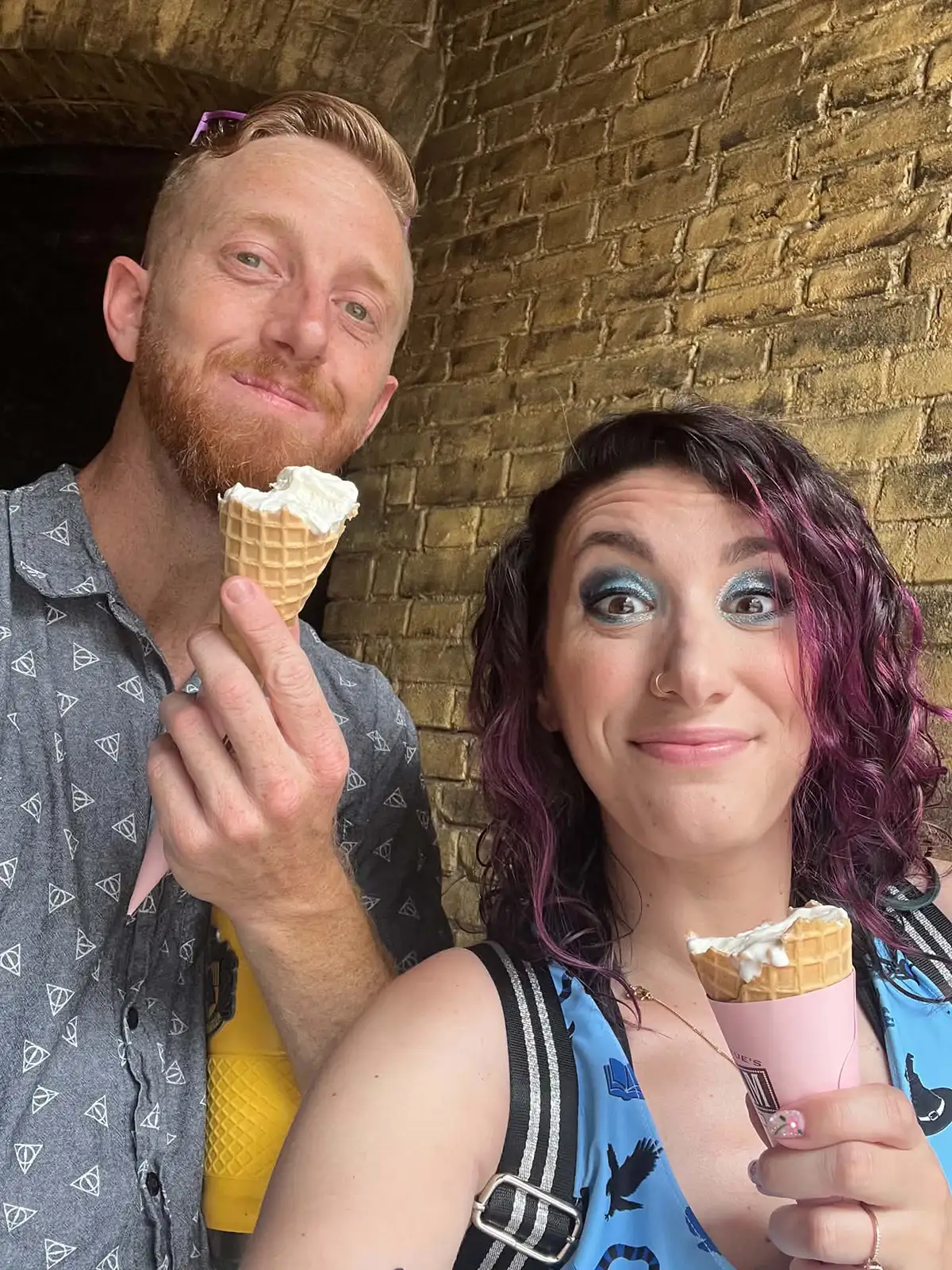 Jessica Thompson and her husband are smiling and holding ice cream cones as they stand in front of a brick wall at The Wizarding World of Harry Potter. The man has short red hair and a beard, while Jessica’s wavy purple hair and bold blue eye makeup add a touch of magic to the scene. - Favorite Grampy Travels