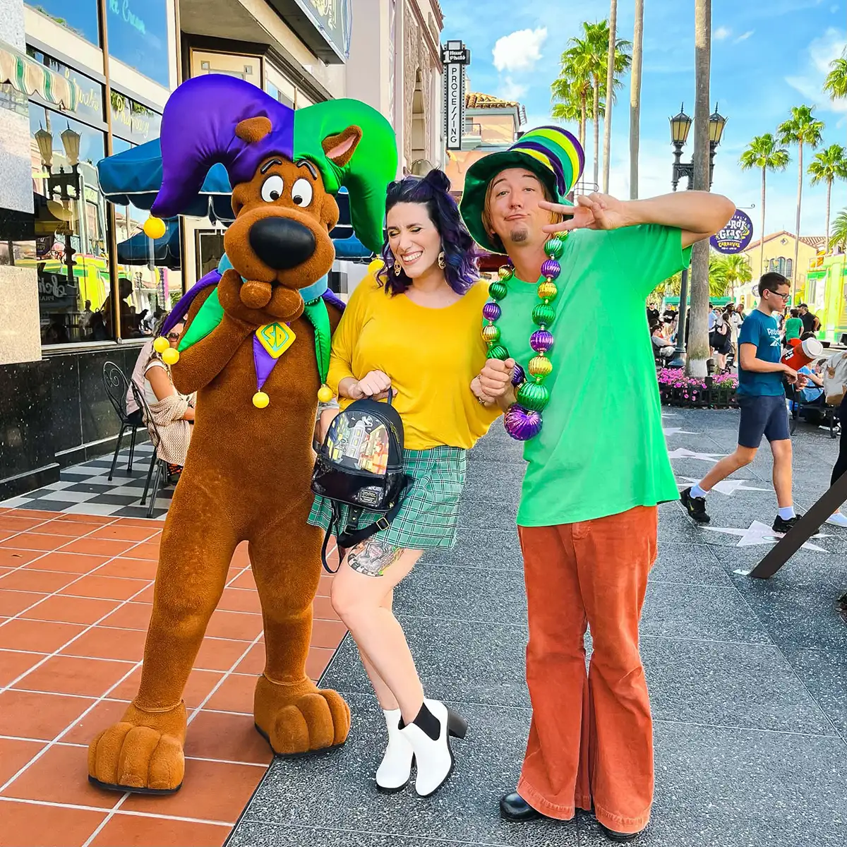 Three people pose together outdoors: one in a Scooby-Doo costume, a woman in a yellow top and green skirt, and a man in a green shirt with colorful bead necklaces and a jester hat. Palm trees and shops are in the background. - Favorite Grampy Travels