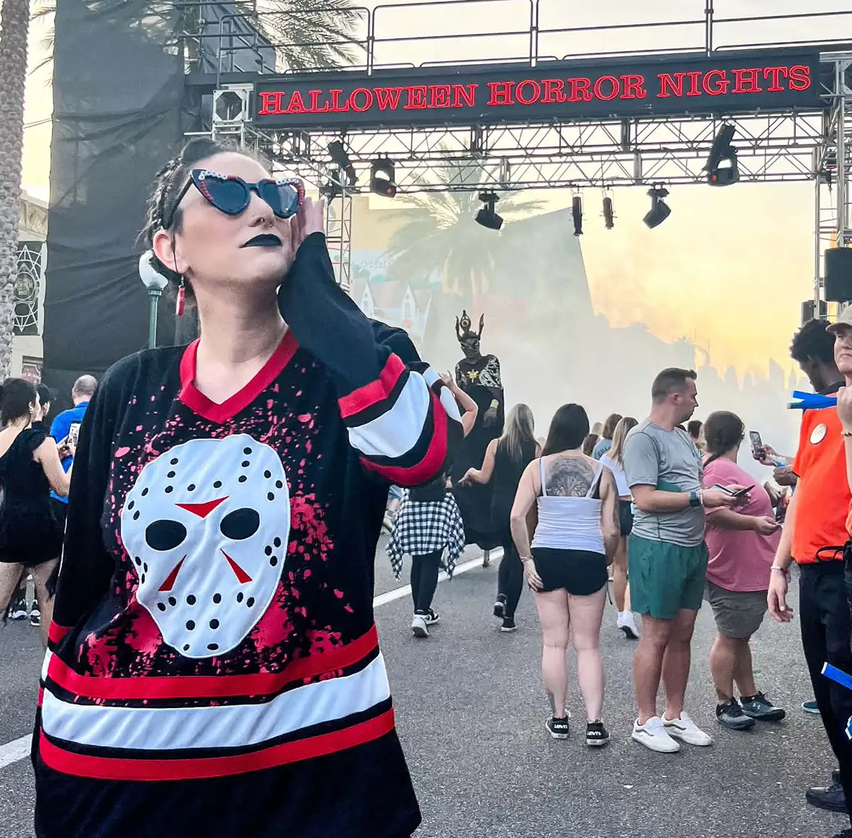 A person in a Jason hockey mask shirt and sunglasses stands at Halloween Horror Nights, with crowds and spooky decorations in the background. - Favorite Grampy Travels