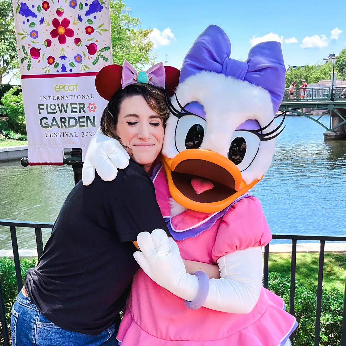 A woman wearing Minnie Mouse ears hugs Daisy Duck in a pink dress at Epcots International Flower & Garden Festival, with a river and festival sign in the background. - Favorite Grampy Travels