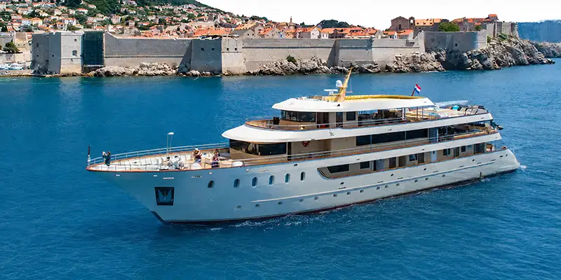 the Lastacica , a Large White Yacht, Sails on Blue Water Near a Historic Walled City with Orange-roofed Buildings and Rocky Shores in the Background. - Favorite Grampy Travels - Lastacica Yacht Cruising - Favorite Grampy Travels 2026 - Favorite Grampy Travels 2026