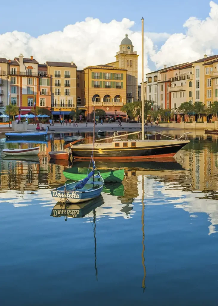 Colorful Boats Float on Calm Water in a Harbor, Reflecting Nearby Pastel Buildings and a Clock Tower Under a Partly Cloudy, Blue Sky. the Scene Feels Peaceful and Inviting, Evoking a Mediterranean or European Coastal Town but It is Portofino Bay Hotel at Universal Orlando Resort. - Favorite Grampy Travels - Favorite Grampy Travels 2026 - Favorite Grampy Travels 2026
