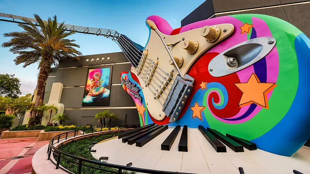 a Giant, Colorful Electric Guitar Sculpture with Swirling Designs and Stars Sits Outdoors Near Palm Trees and a Mural on a Building Wall, with a Roller Coaster Track Overhead. - Favorite Grampy Travels - Favorite Grampy Travels 2026