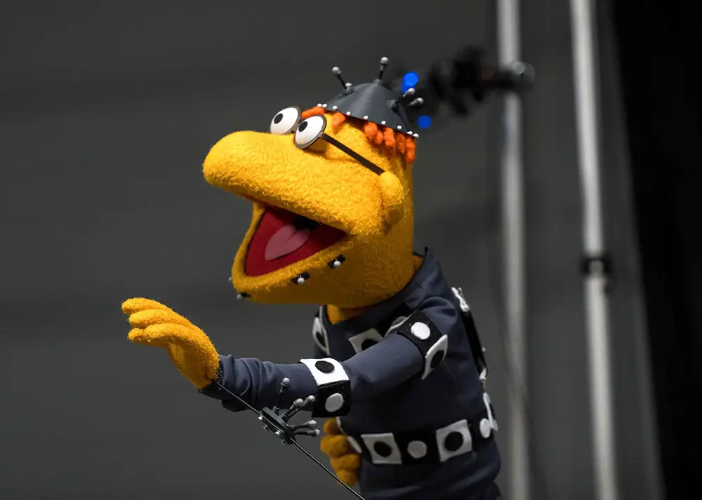 a Yellow Muppet in a Motion Capture Suit with Sensors and a Helmet, Standing with Its Mouth Open and Arm Raised, Against a Blurred Background with Metal Poles. - Favorite Grampy Travels - Favorite Grampy Travels 2026