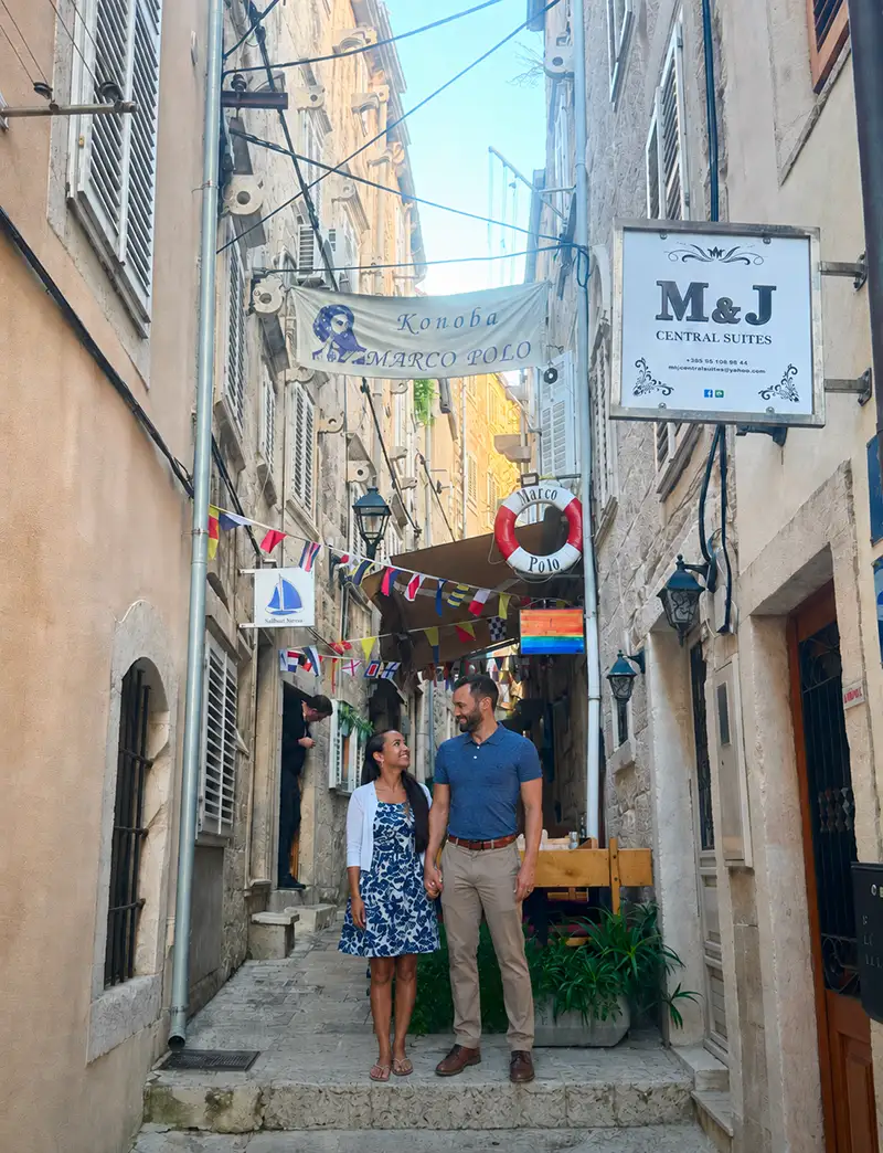 a Couple Stands Smiling Together in a Narrow, Colorful Alley Lined with Flags, Shutters, and Signs, Including M&j Central Suites and Marco Polo. the Setting Appears Lively and European. - Favorite Grampy Travels - Favorite Grampy Travels 2026 - Favorite Grampy Travels 2026