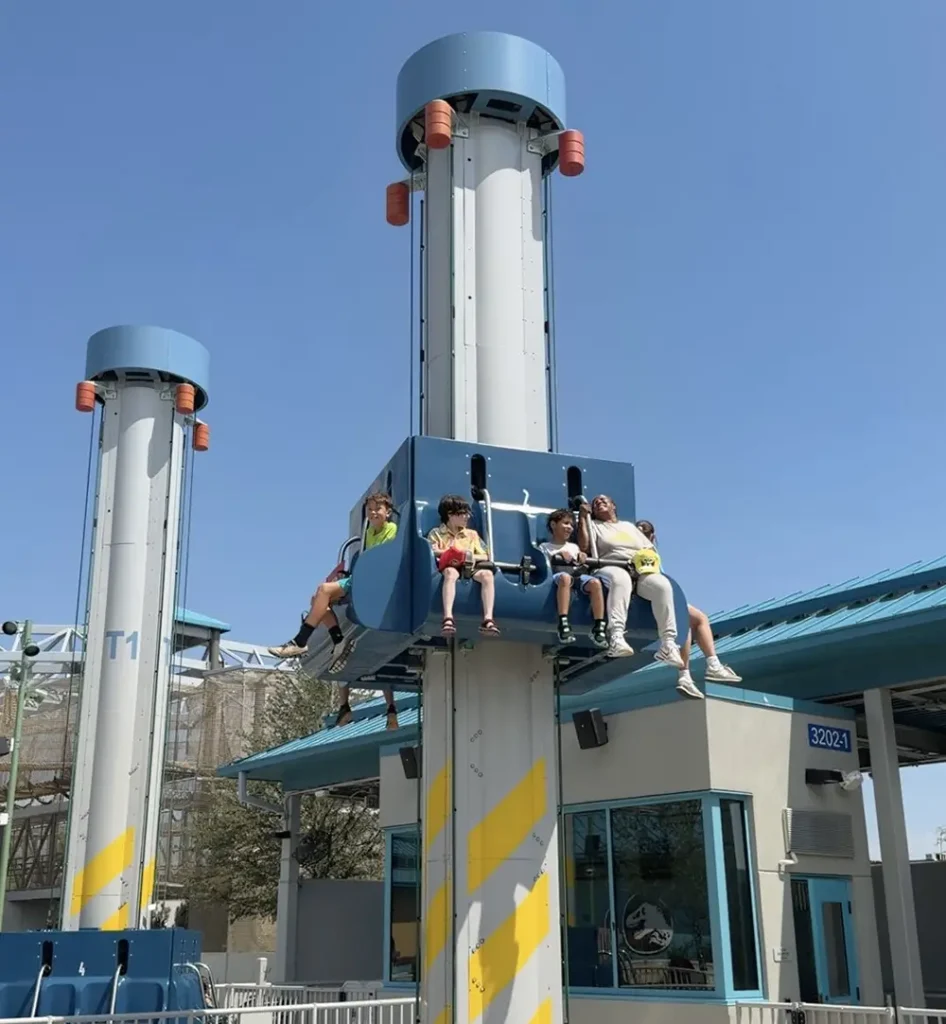 Ukr Jurassic World Pteranodrop - Favorite Grampy Travels Five people sit on an amusement park drop ride in mid-air, some with legs dangling, against a clear blue sky and a building with blue trim in the background. - Favorite Grampy Travels