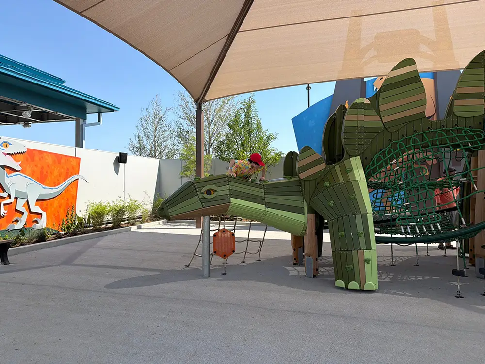 Ukr-jurassic-world-paddock-play - Favorite Grampy Travels A child in a red hat plays on a large green dinosaur-shaped playground structure under a shade canopy, with dinosaur murals on the nearby wall. - Favorite Grampy Travels