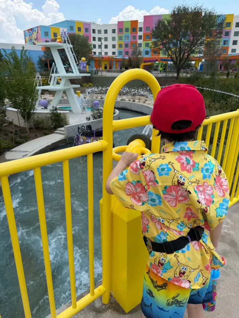 Ukr-minions-bello-bay-cruise-water-launcher - Favorite Grampy Travels A child in a red cap and colorful shirt looks through a viewer on a yellow bridge, with a pool, diving boards, and a colorful building in the background. - Favorite Grampy Travels