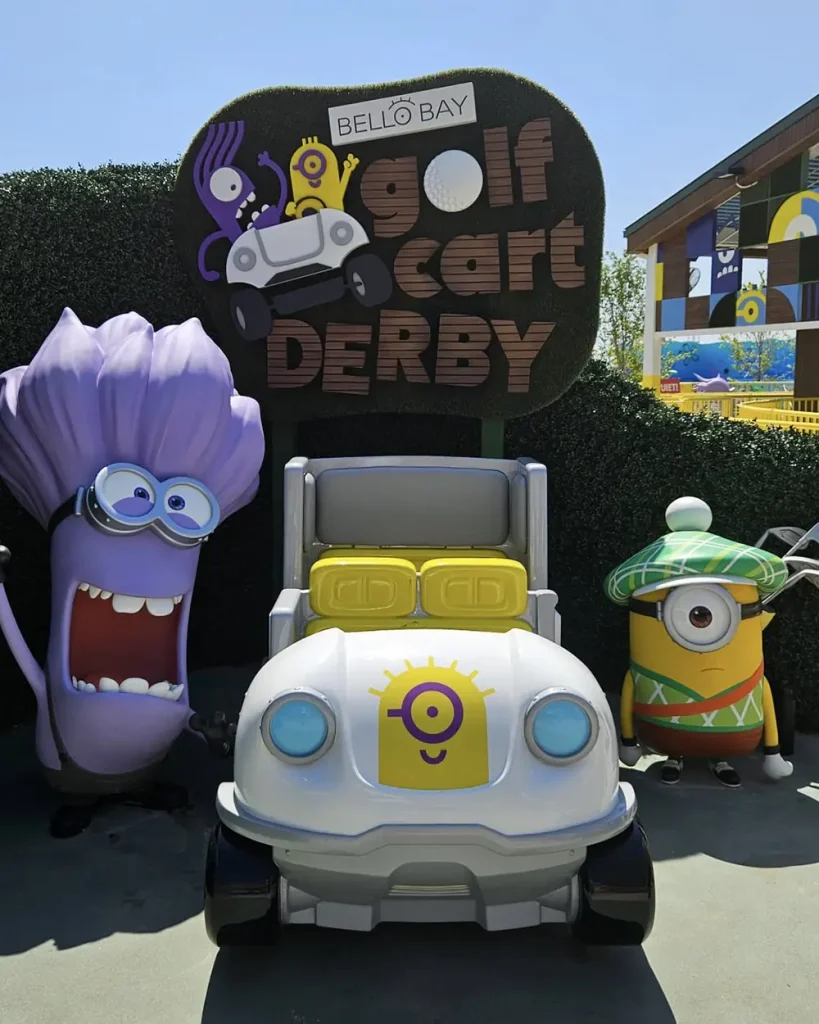 Ukr-minions-bello-bay-golf-cart-derby-statue - Favorite Grampy Travels Two Minion characters in costumes stand beside a white golf cart with a Minion logo, under a sign that reads Bello Bay Golf Cart Derby. Greenery and buildings are visible in the background. - Favorite Grampy Travels
