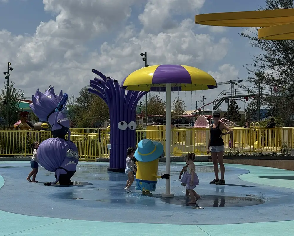 Ukr-minions-bello-bay-water-play - Favorite Grampy Travels Children play in a splash pad area with Minion-themed water features, including purple and yellow statues and an umbrella fountain. An adult stands nearby under a partly cloudy sky. - Favorite Grampy Travels