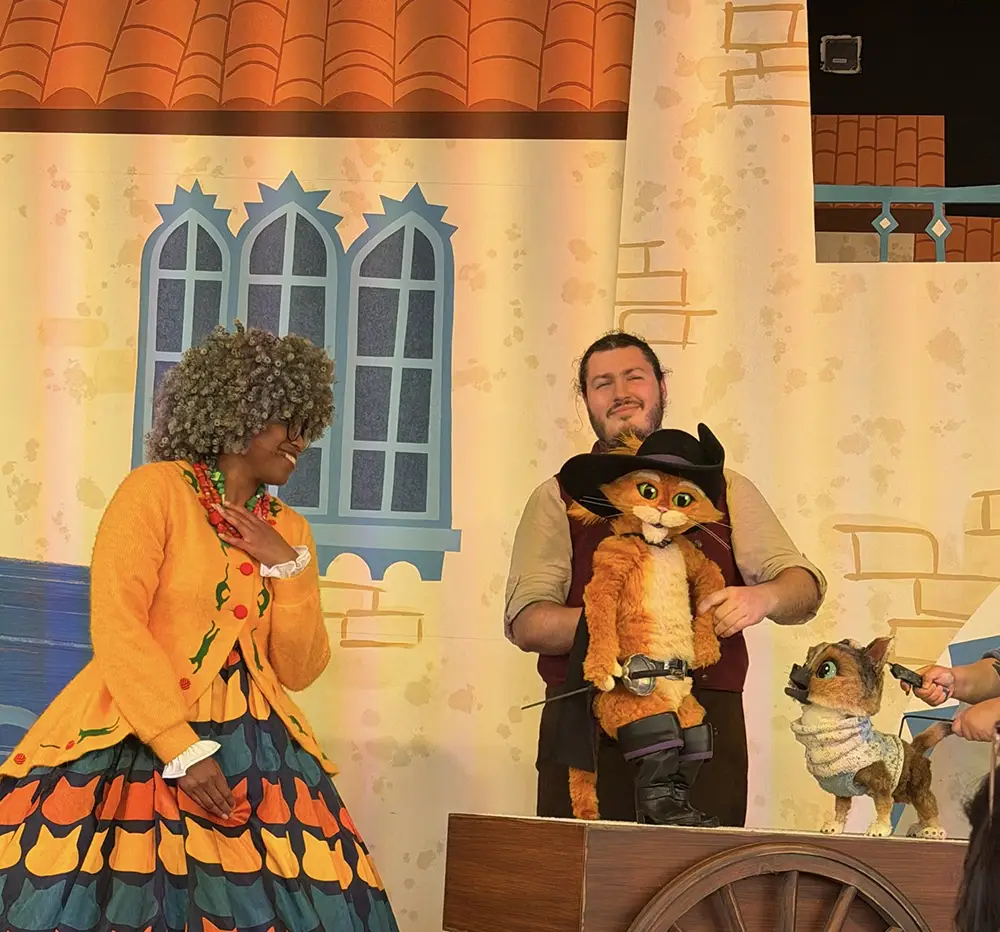 Ukr-puss-in-boots-del-mar-mama-lunas-adopt-a-kitty-day-puppets - Favorite Grampy Travels Two people in colorful costumes stand onstage with puppets of Puss in Boots and a dog, set against a backdrop of a building with blue windows and a tiled roof. - Favorite Grampy Travels