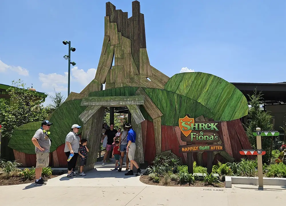 Ukr-shrek-and-fionas-happily-ogre-after - Favorite Grampy Travels A group of people stands outside the entrance to Shrek & Fionas Happily Ever After, which features a colorful, wooden tree design and a bright sign. Its a sunny day with a clear blue sky. - Favorite Grampy Travels