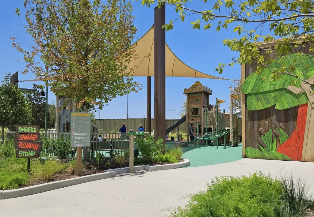 Ukr-shreks-swamp-rompin-stomp - Favorite Grampy Travels A colorful outdoor playground features a wooden playhouse, green play area, trees, and a sign reading Shreks Swamp Roarin Stomp. Bright shade sails cover parts of the park under a clear blue sky. - Favorite Grampy Travels