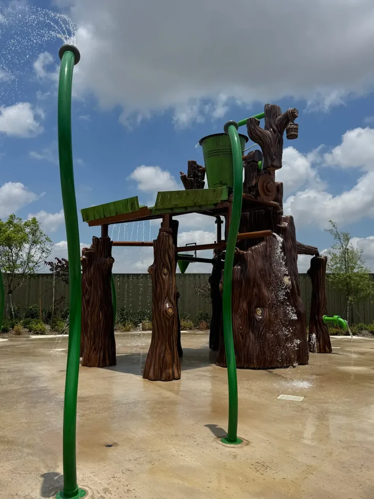 Ukr-shreks-swamp-splash - Favorite Grampy Travels A wooden-themed splash pad structure with a big green bucket on top, spraying and dumping water, surrounded by green poles spraying water, under a partly cloudy sky. - Favorite Grampy Travels