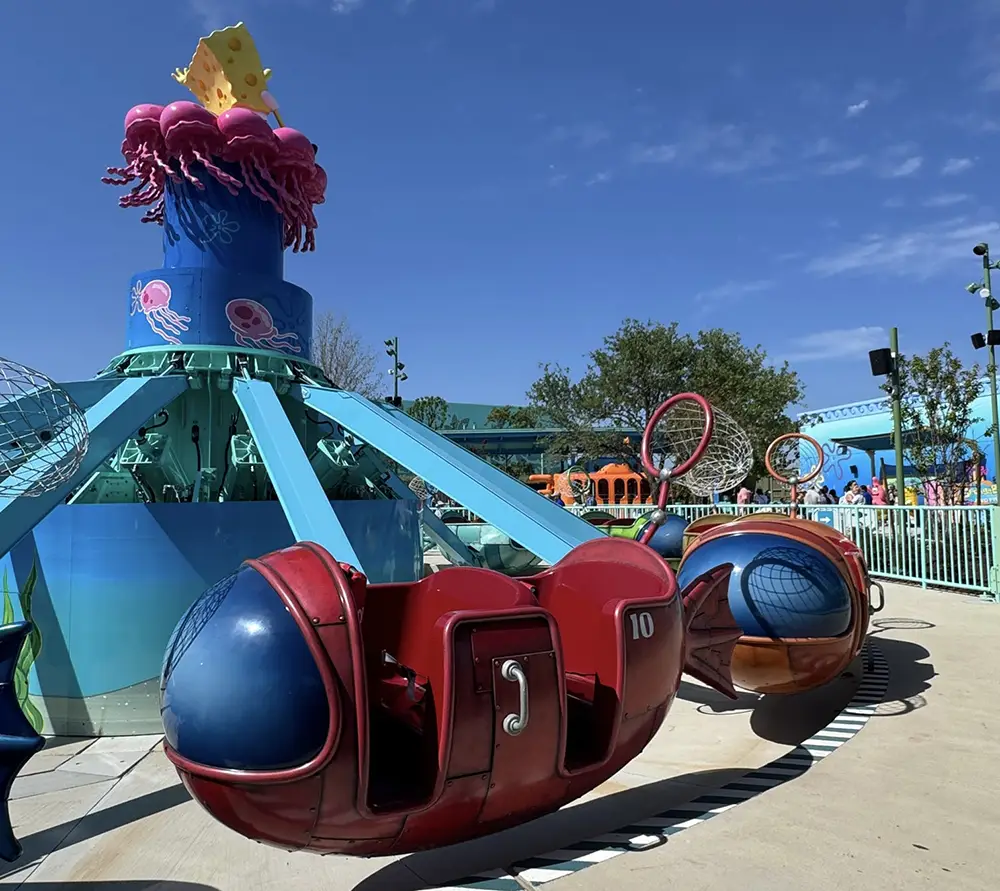 Ukr-spongbob-squarepants-bikini-bottom-jellyfish-fields-jamboree - Favorite Grampy Travels A colorful amusement park ride with red and blue submarine-shaped seats, surrounded by teal beams, sits under a blue sky. The ride has jellyfish and sea-themed decorations on top, creating a vibrant sensory experience for visitors. - Favorite Grampy Travels