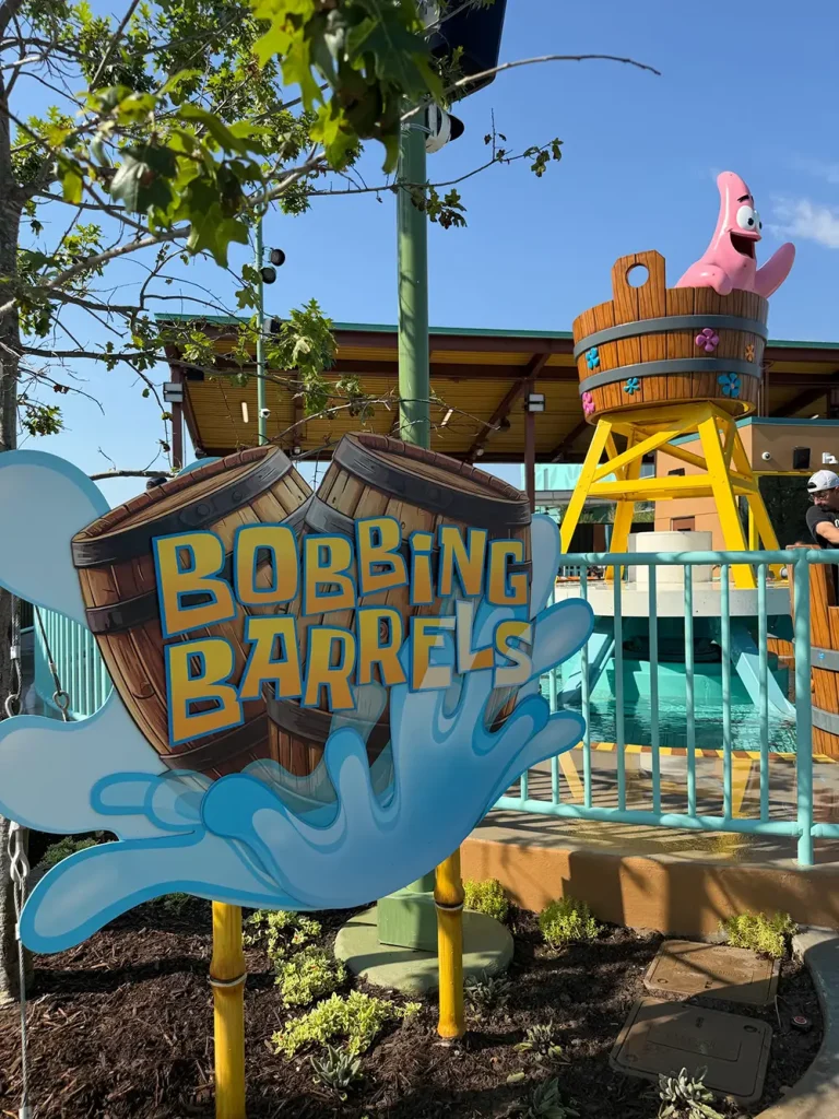 Ukr-spongebob-squarepants-bikini-bottom-bobbing-barrels-sign - Favorite Grampy Travels Sign for Bobbing Barrels with water splash graphics evokes a sensory experience. Behind it is a water ride featuring a large barrel and a pink cartoon starfish character perched on top. Lush plants and a blue fence complete the lively background. - Favorite Grampy Travels
