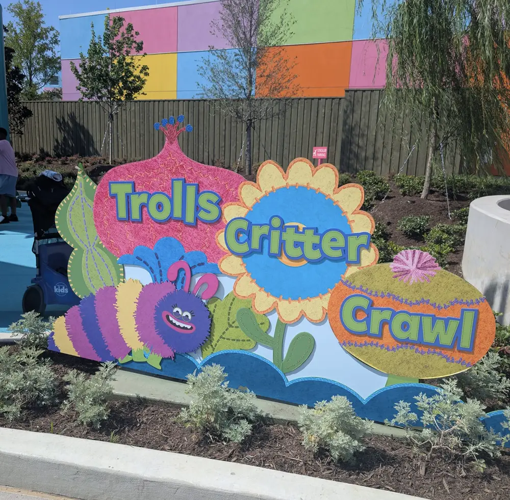 Ukr-trolls-critter-crawl-sign - Favorite Grampy Travels Colorful sign reading Trolls Critter Crawl with cartoon flowers and a smiling, striped caterpillar, set in a garden near a multicolored building. - Favorite Grampy Travels