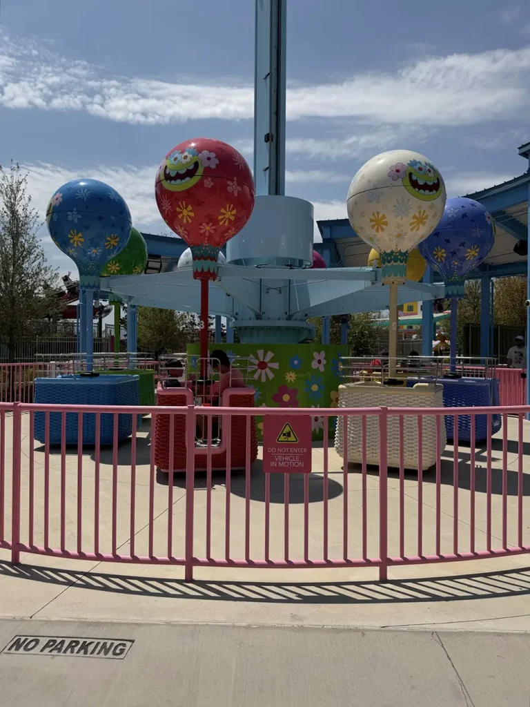Ukr-trolls-hair-in-the-clouds - Favorite Grampy Travels A colorful amusement park ride with hot air balloon-themed seats, surrounded by a pink fence, stands empty under a sunny sky. No Parking is painted on the curb in front. - Favorite Grampy Travels