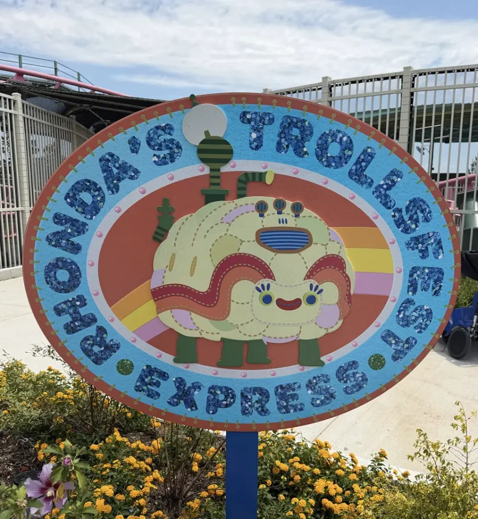 Ukr-trolls-rhondas-trollfest-express-sign - Favorite Grampy Travels Colorful sign reading Rhondas Trollsfest Express with a cartoon creature and rainbow, surrounded by flowers, near a metal fence and blue sky. - Favorite Grampy Travels