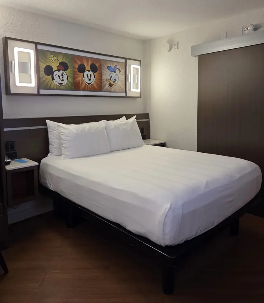 a Neatly Made Bed in a Hotel Room with White Linens, Flanked by Nightstands. Above the Bed is Artwork Featuring Cartoon Faces of Minnie Mouse, Mickey Mouse, and Donald Duck. - Favorite Grampy Travels - Favorite Grampy Travels 2026