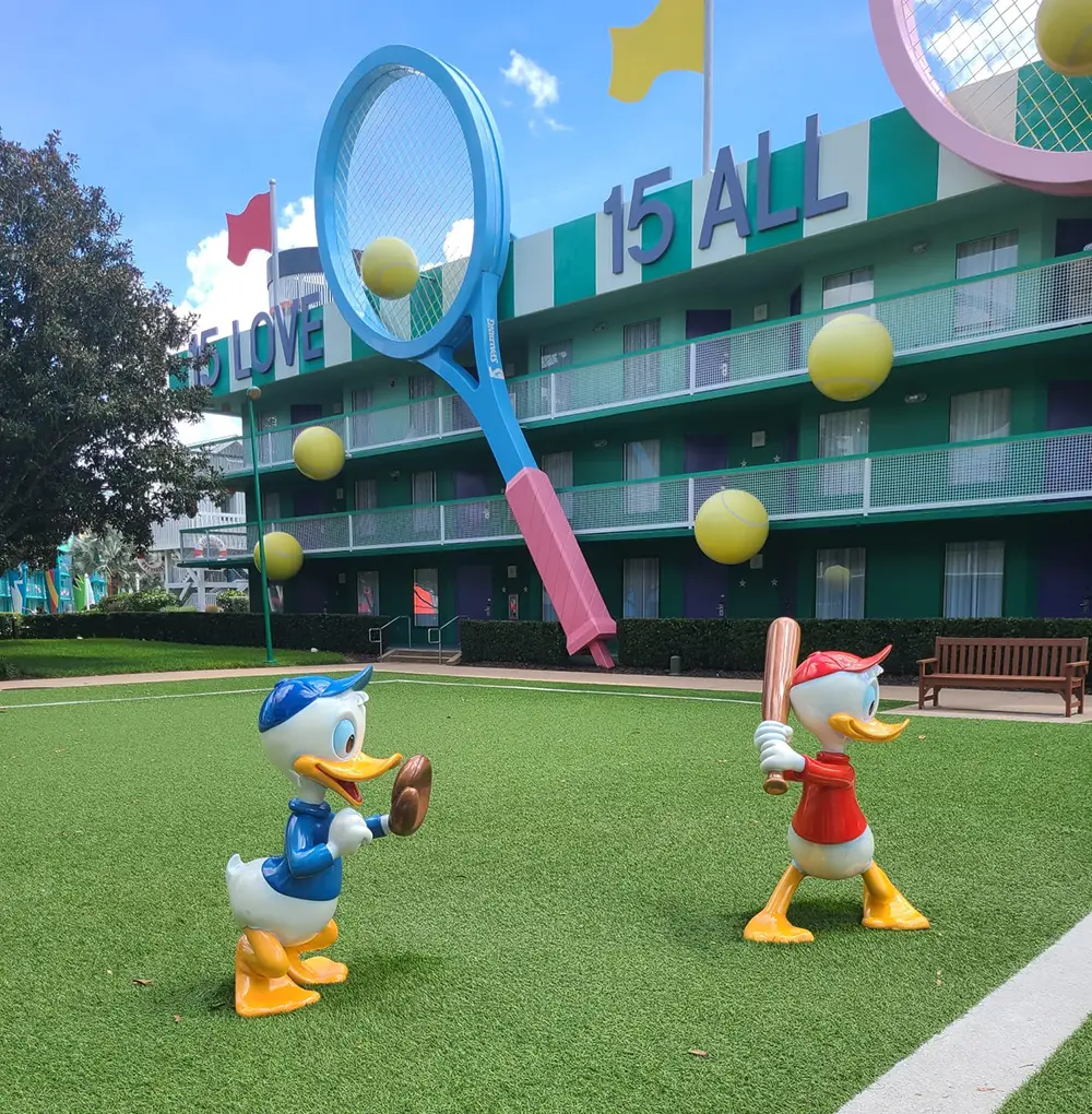 Two Cartoon Duck Statues Play Baseball on Green Turf Near a Building with Tennis-themed Decor, Oversized Tennis Rackets, and Floating Tennis Balls. the Building Displays the Phrases 15 Love and 15 All.. - Favorite Grampy Travels - Favorite Grampy Travels 2026