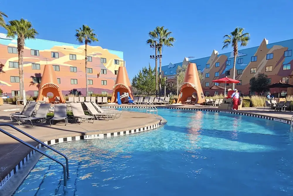 Hotel pool with lounge chairs, orange cone-shaped cabanas, palm trees, a lifeguard on duty, and a colorful building with painted designs in the background under a clear blue sky. - Favorite Grampy Travels