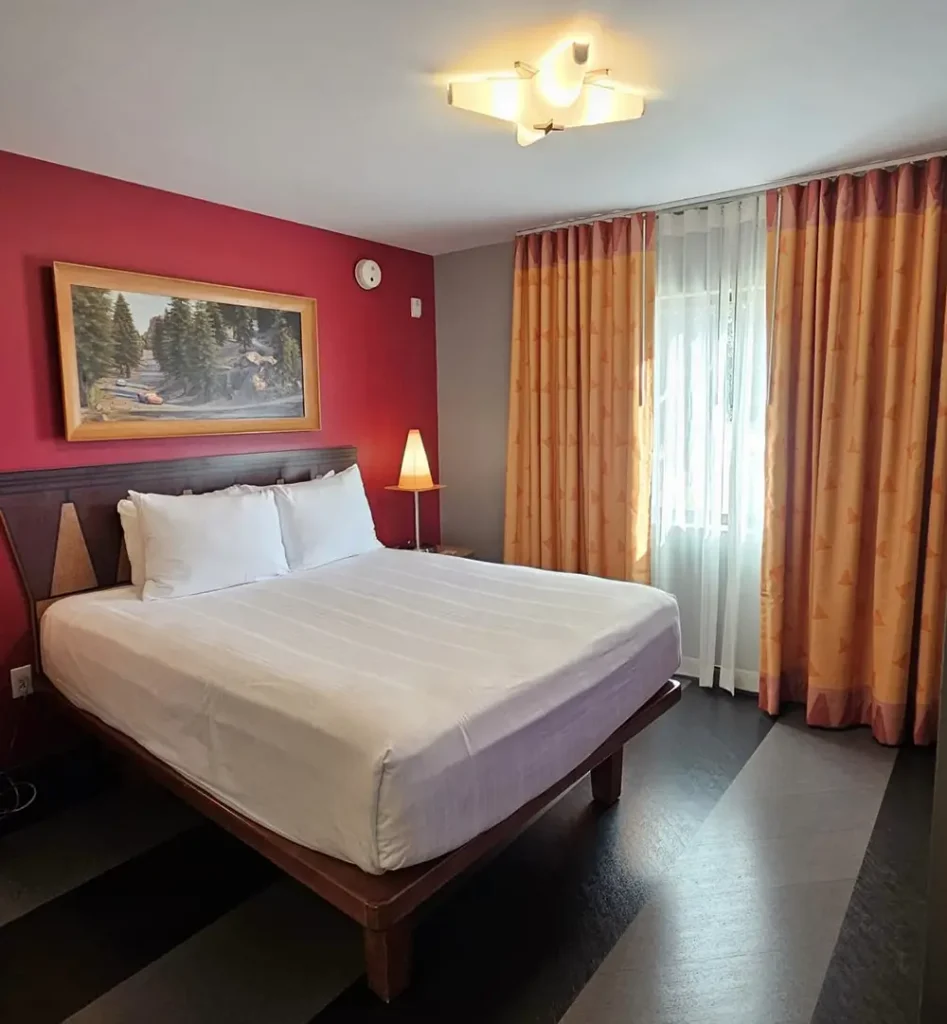 Wdw-art-of-animation-cars-room - Favorite Grampy Travels a Neatly Made Bed in a Hotel Room with Red and Beige Walls, a Painting Above the Headboard, Orange Curtains, a Lamp, and Sunlight Streaming Through a Window with Sheer White Curtains. - Favorite Grampy Travels - Favorite Grampy Travels 2026