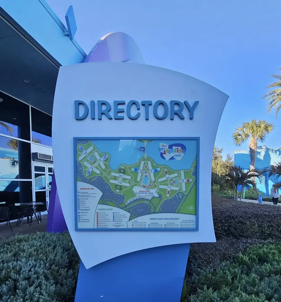 Wdw-art-of-animation-directory - Favorite Grampy Travels Outdoor Directory Sign with a Colorful Site Map and Labeled Locations, Placed Beside a Modern Building with Blue Walls, Some Greenery, and Palm Trees in the Background Under a Clear Sky. - Favorite Grampy Travels - Favorite Grampy Travels 2026