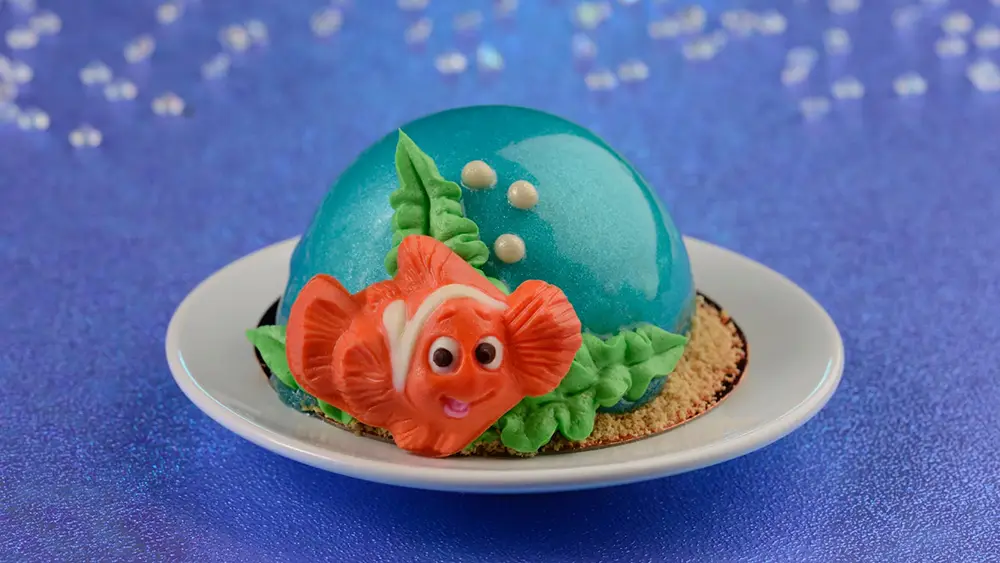 Wdw-art-of-animation-nemo-dessert - Favorite Grampy Travels a Dome-shaped Blue Dessert with a Shiny Glaze, Decorated with an Orange Fish Made of Icing and Green Leaves, Sits on a White Plate Against a Sparkling Purple Background. - Favorite Grampy Travels - Favorite Grampy Travels 2026
