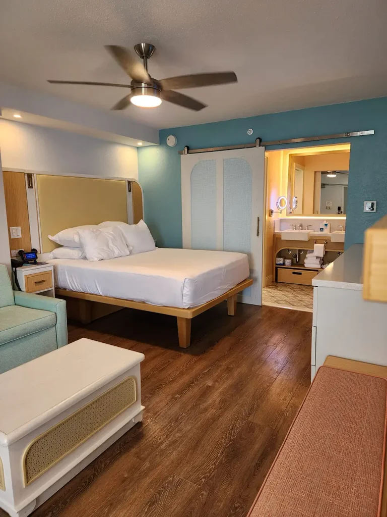 A modern hotel room with a queen bed, ceiling fan, mint green chair, ottoman, wooden floors, and an open sliding door showing a well-lit bathroom with a mirror and sink. - Favorite Grampy Travels