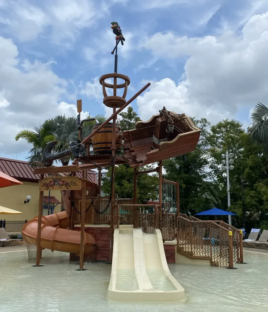 Pirate ship-themed water play structure with slides, a lookout, and a parrot figure on top, surrounded by shallow water, trees, and a partly cloudy sky. - Favorite Grampy Travels