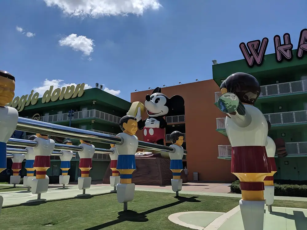 Wdw-pop-century-resort-courtyard-foosball - Favorite Grampy Travels Large Foosball Player Figures Stand in Front of a Colorful Hotel Building, with a Giant Mickey Mouse Statue in the Background and a Bright Blue Sky Overhead. - Favorite Grampy Travels - Favorite Grampy Travels 2026