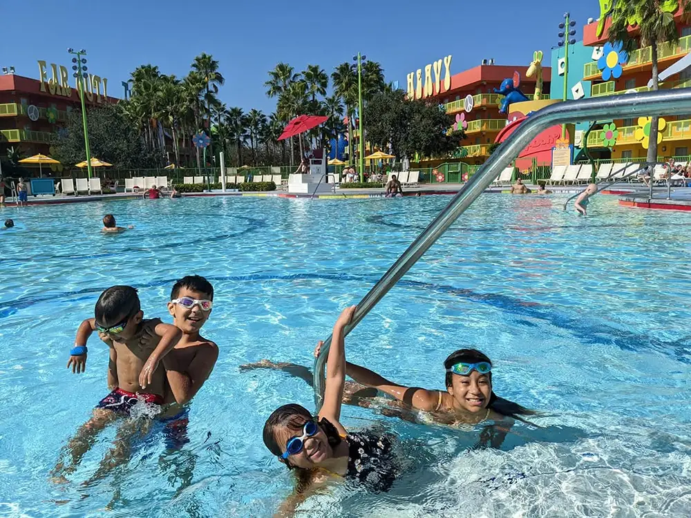 Wdw-pop-century-resort-kids-in-pool - Favorite Grampy Travels Four Kids in Swimsuits and Goggles Play Together in a Bright, Outdoor Pool with Colorful Buildings and Palm Trees in the Background. One Child Holds a Pool Railing While Others Smile and Splash Nearby. - Favorite Grampy Travels - Favorite Grampy Travels 2026