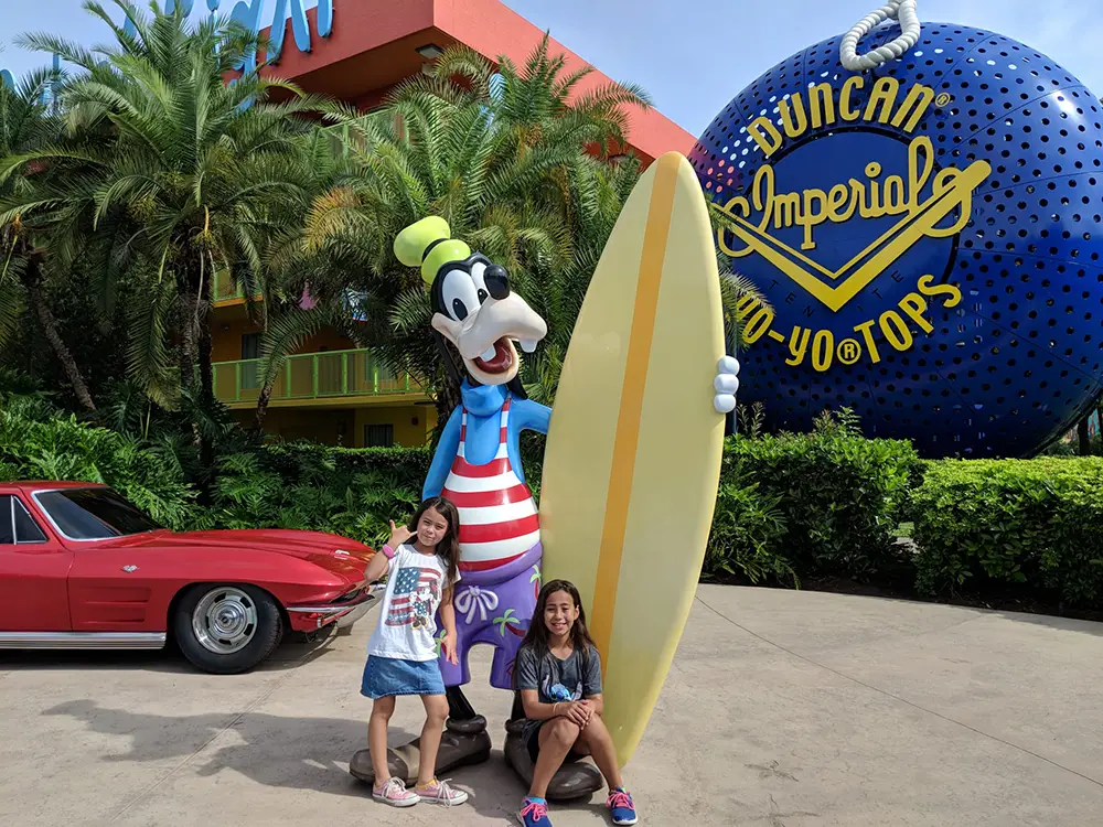 Wdw-pop-century-resort-kids-with-surfer-goofy - Favorite Grampy Travels Two Girls Pose and Smile in Front of a Tall Goofy Statue Holding a Surfboard, with a Red Car and a Giant Blue Yo-yo Sculpture in the Background, Surrounded by Palm Trees and Greenery. - Favorite Grampy Travels - Favorite Grampy Travels 2026