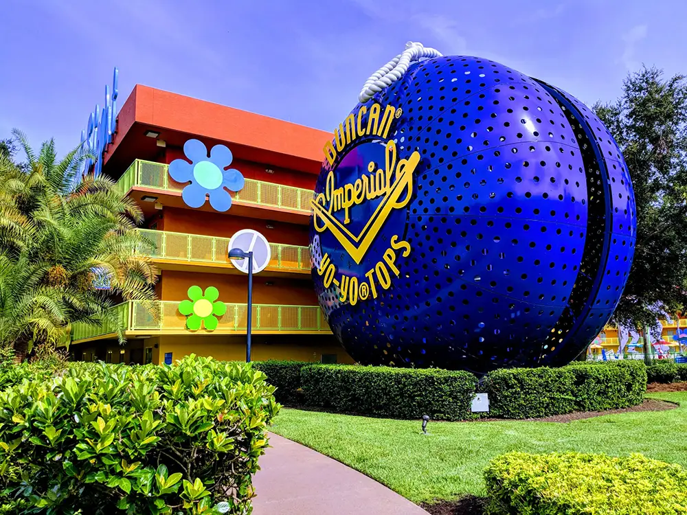 A large blue Duncan yo-yo sculpture stands beside a colorful hotel building decorated with oversized flowers and yellow railings, surrounded by green bushes and trees under a blue sky. - Favorite Grampy Travels