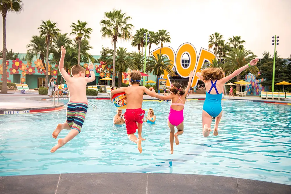 Wdw-pop-century-resort-pool-kids-jumping-in - Favorite Grampy Travels Four Children in Swimsuits Jump into a Pool While Two Adults Watch from the Water. Palm Trees and Colorful Decorations, Including Large Pop Letters, Are in the Background on a Sunny Day. - Favorite Grampy Travels - Favorite Grampy Travels 2026