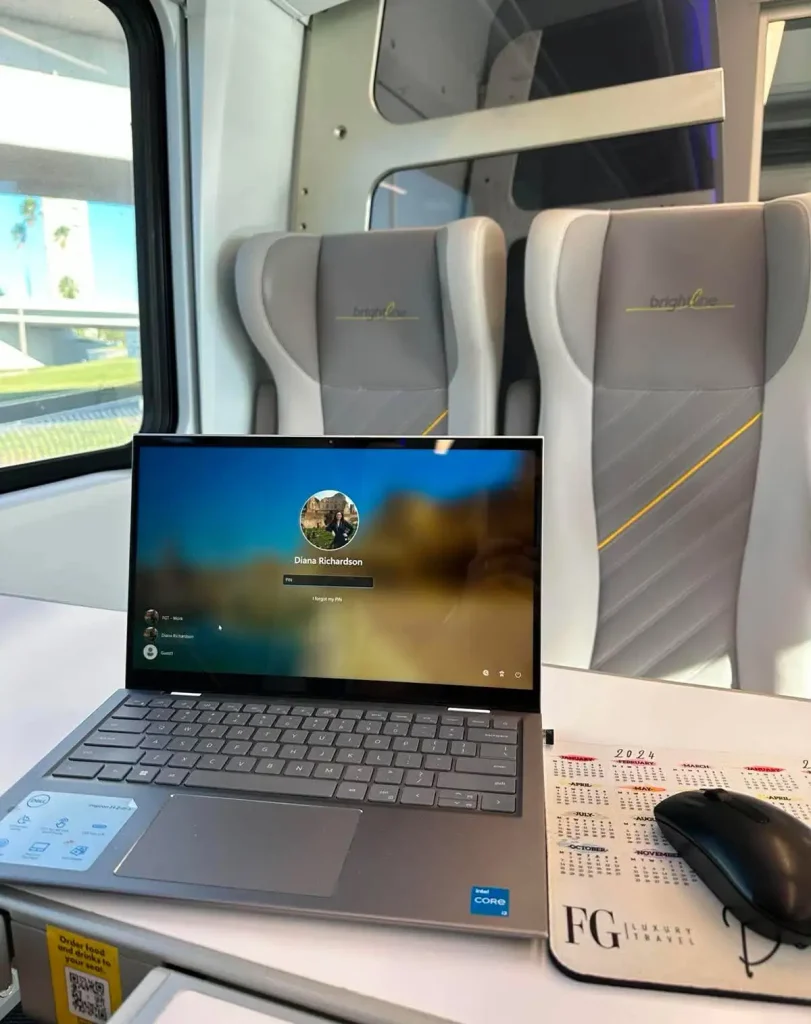 A laptop on a tray table inside a modern train, with a wireless mouse and a 2023 calendar mousepad; gray seats and a window are visible in the background. - Favorite Grampy Travels