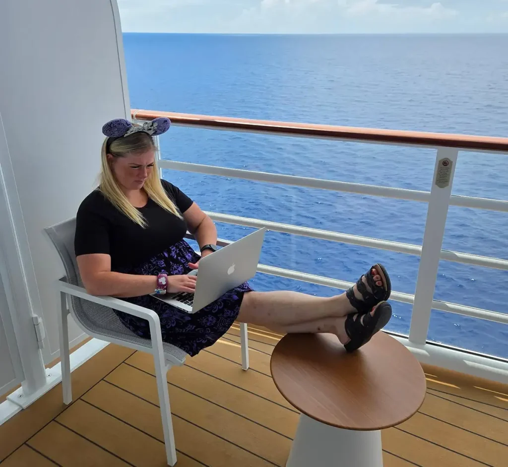 A Travel Agent wearing Mickey Mouse ears sits on a chair on a cruise ship balcony, working on a laptop helping clients with her feet propped up on a small table, ocean in the background. - Favorite Grampy Travels