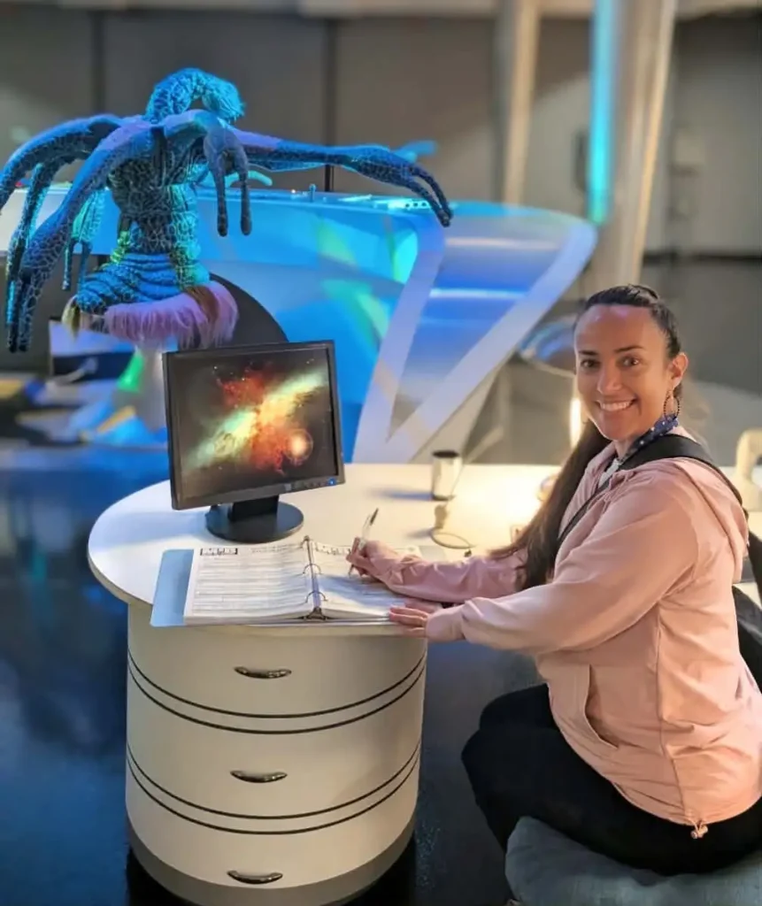 Nikki Hendrix writes in a binder at a futuristic white desk with a monitor showing a galaxy at the Men in Black attraction at Universal Studios in Orlando. Behind her is a blue, tentacled alien figure on display in a well-lit, modern room. - Favorite Grampy Travels