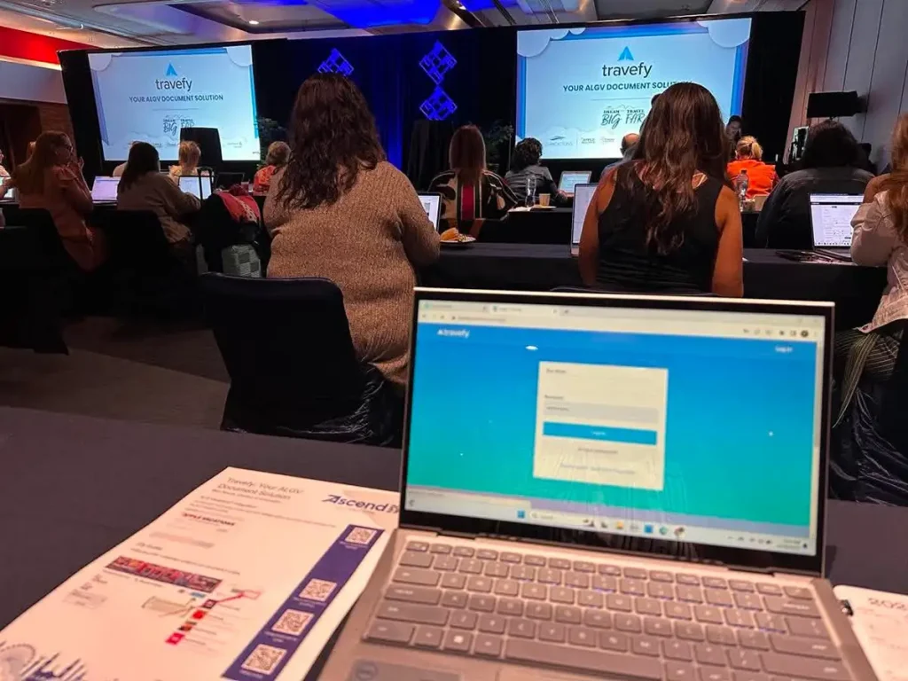 People attend a conference in a dimly lit room, watching a presentation on a large screen. A laptop with the Travefy login page is open in the foreground. Handouts and notes rest on the table beside the laptop. - Favorite Grampy Travels